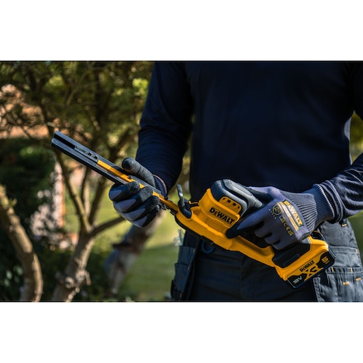 A person wearing gloves and dark clothing is holding a yellow DEWALT cordless hedge trimmer outdoors, with trees in the background.