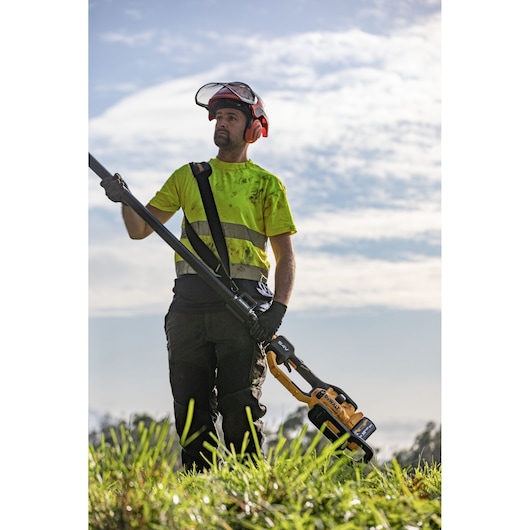 A person wearing safety gear and a high-visibility shirt stands outdoors on grass, holding a DEWALT DCMPS640N pole saw. The sky is partly cloudy in the background.