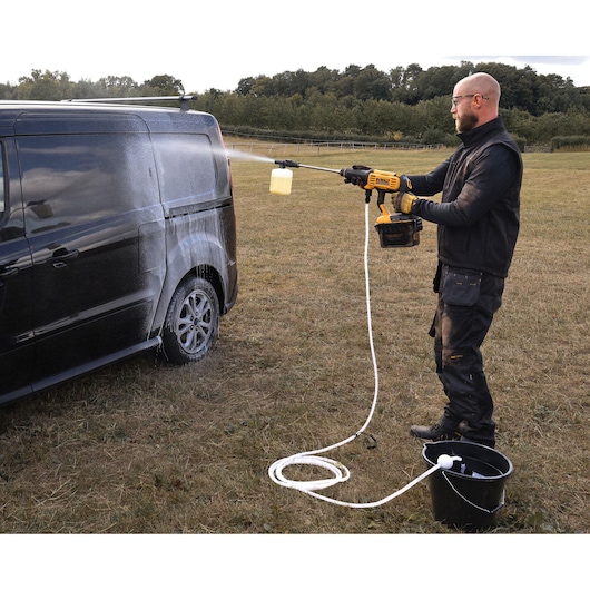 Person using a DEWALT cordless pressure washer (model DCMPW1000N) to wash a black van outdoors. The washer is connected via a hose to a bucket of water and sprays foam onto the vehicle. The person is wearing work clothes and gloves in a grassy field.