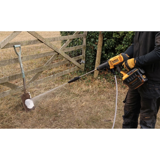 A person is using a DEWALT portable pressure washer to clean a dirty shovel outdoors near a wooden gate.