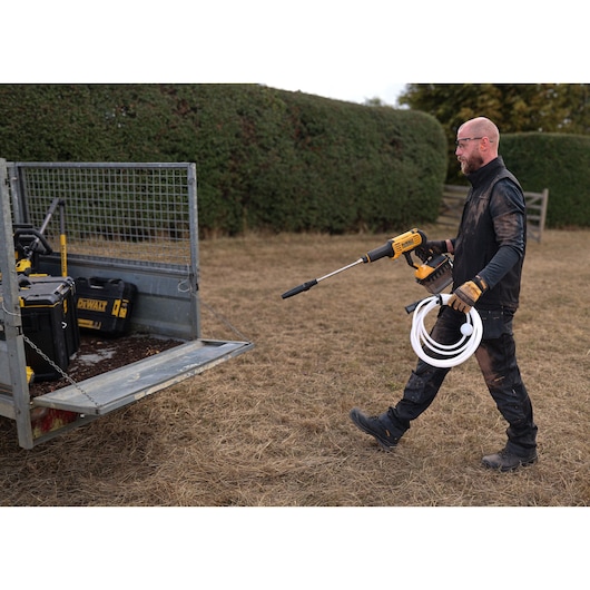 A person wearing work clothes and gloves is carrying a DEWALT portable power washer with a hose, walking near a metal trailer containing DEWALT toolboxes, outdoors on dry grass.