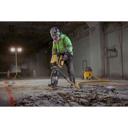 A construction worker in safety gear, including a helmet, gloves, high-visibility jacket, and respiratory mask, operates a yellow industrial jackhammer to break up a concrete floor in a dimly lit underground area. Debris and rubble surround the worker, and orange safety cones and floodlights are visible in the background.