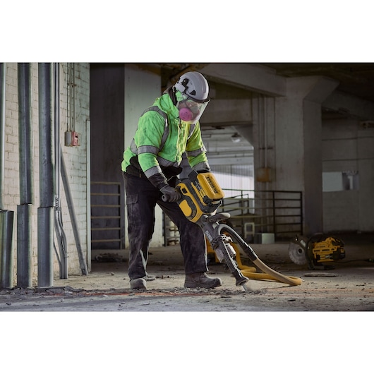 A person wearing safety gear, including a helmet, mask, and reflective jacket, operates a yellow and black industrial jackhammer inside a construction site with concrete floors and exposed pipes.