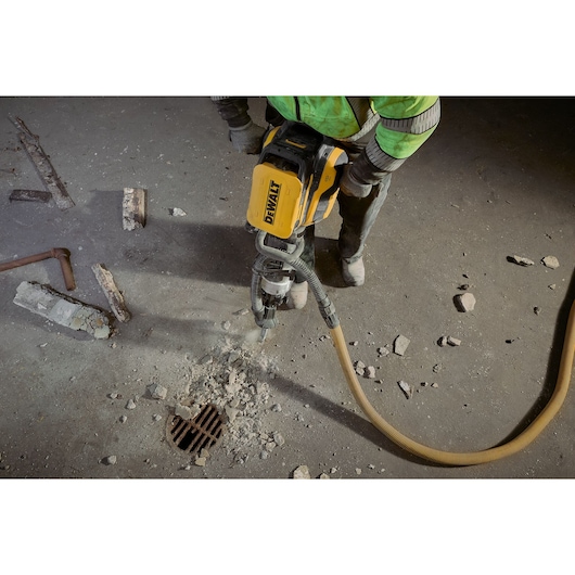 A construction worker using a yellow DEWALT jackhammer to break up concrete near a floor drain, with rubble and debris scattered around.