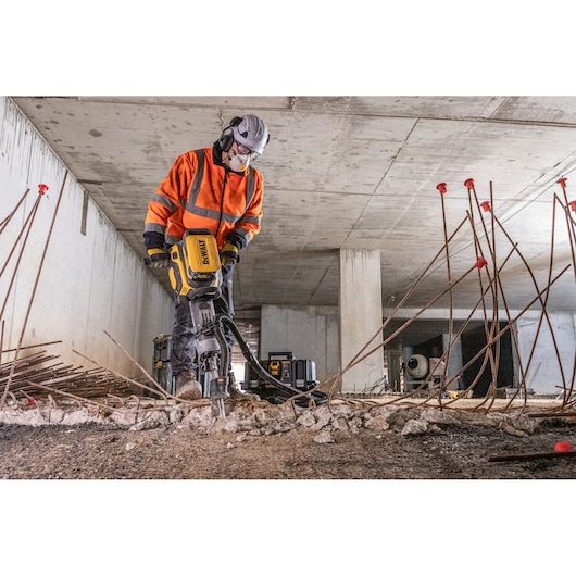 Worker using DEWALT concrete breaker tool indoors with safety gear amid rebar and rubble.