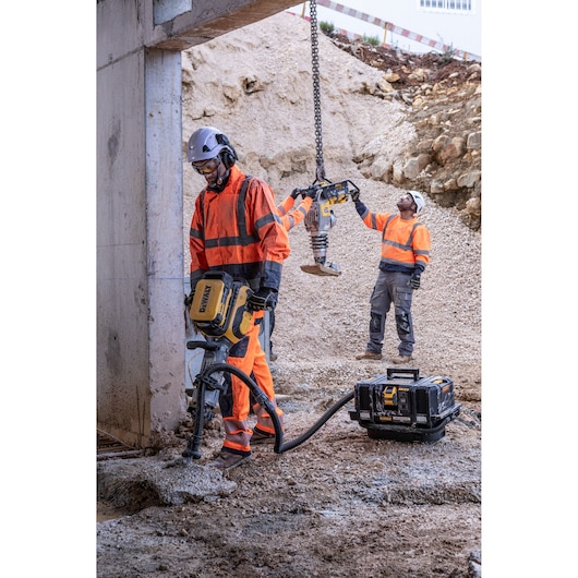 Workers in orange gear use DEWALT tools at a dusty construction site with heavy machinery and a portable power unit.